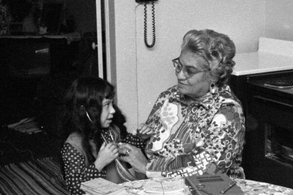 Photo Caption: Lina Chapa Delgado helping her granddaughter Michelle Ortiz listen to her heartbeat in January 1973. On the table are instruments given to Mrs. Delgado by Dr. Hylmar Karbach, Sr., a book on obstetrics from Dr. Frederick Casto and records of some of her 1,600+ deliveries. (New Braunfels Herald negative collection, Feb 1, 1973)