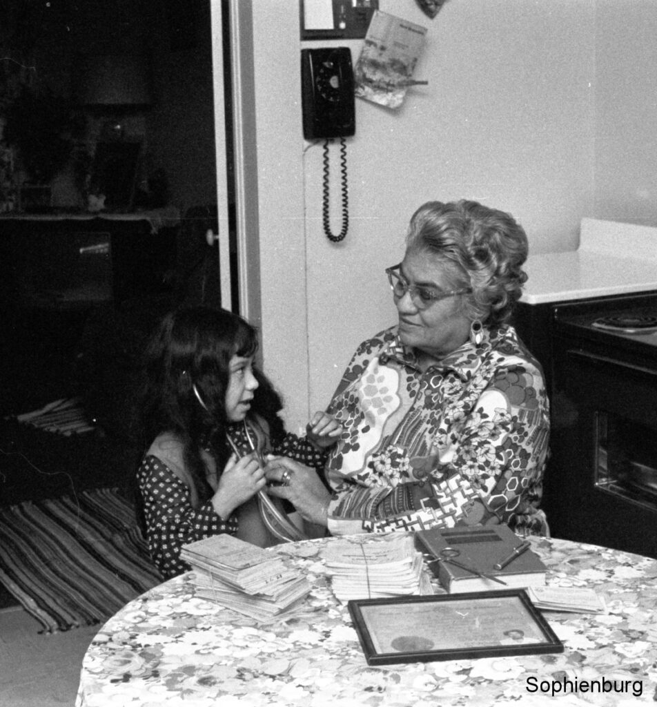 Photo Caption: Lina Chapa Delgado helping her granddaughter Michelle Ortiz listen to her heartbeat in January 1973. On the table are instruments given to Mrs. Delgado by Dr. Hylmar Karbach, Sr., a book on obstetrics from Dr. Frederick Casto and records of some of her 1,600+ deliveries. (New Braunfels Herald negative collection, Feb 1, 1973)
