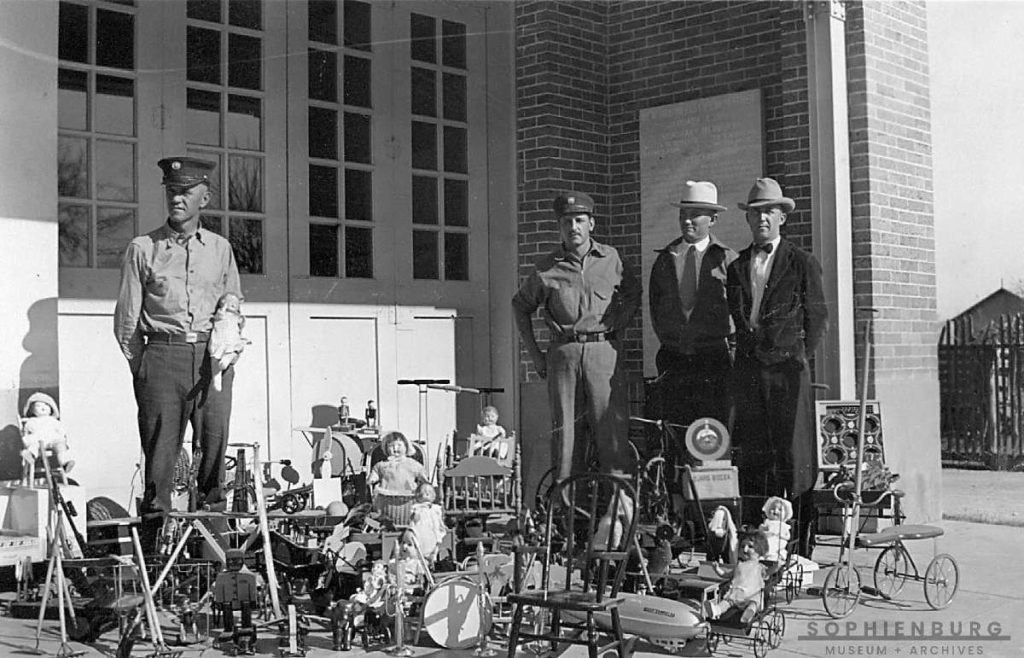 PHOTO CAPTION: L-R, Fireman H.H. Chili Voigt stands with unknown fireman and others (most likely Lions Club members) looking at the toys readied for delivery to needy children, circa 1935.